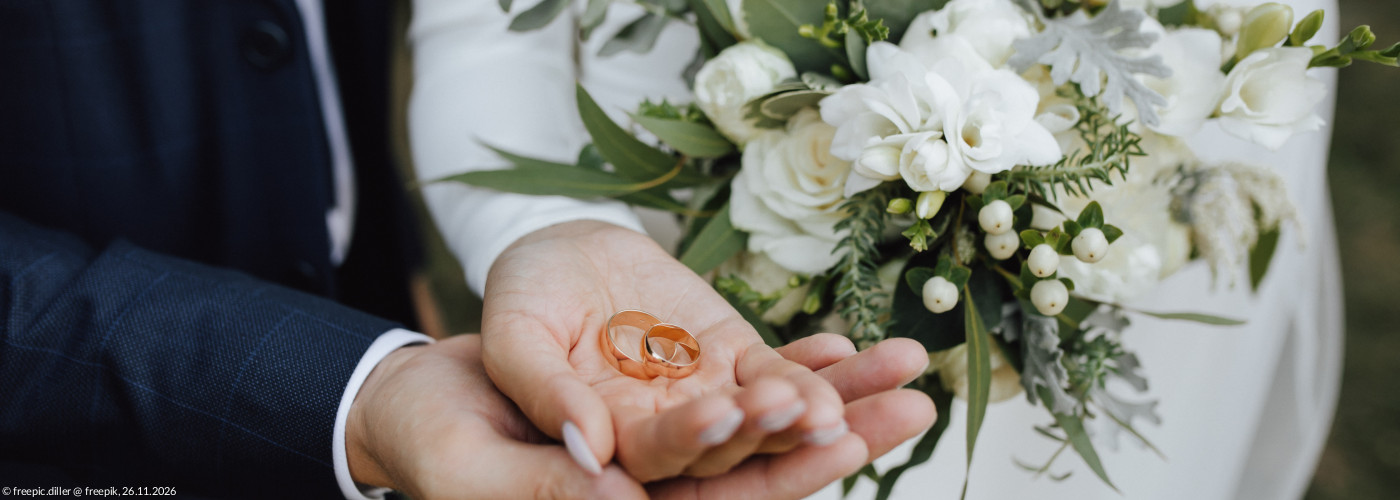 Ein Brautpaar hält Eheringe gemeinsam in der Hand, ein weißer Brautstrauß ist zu sehen