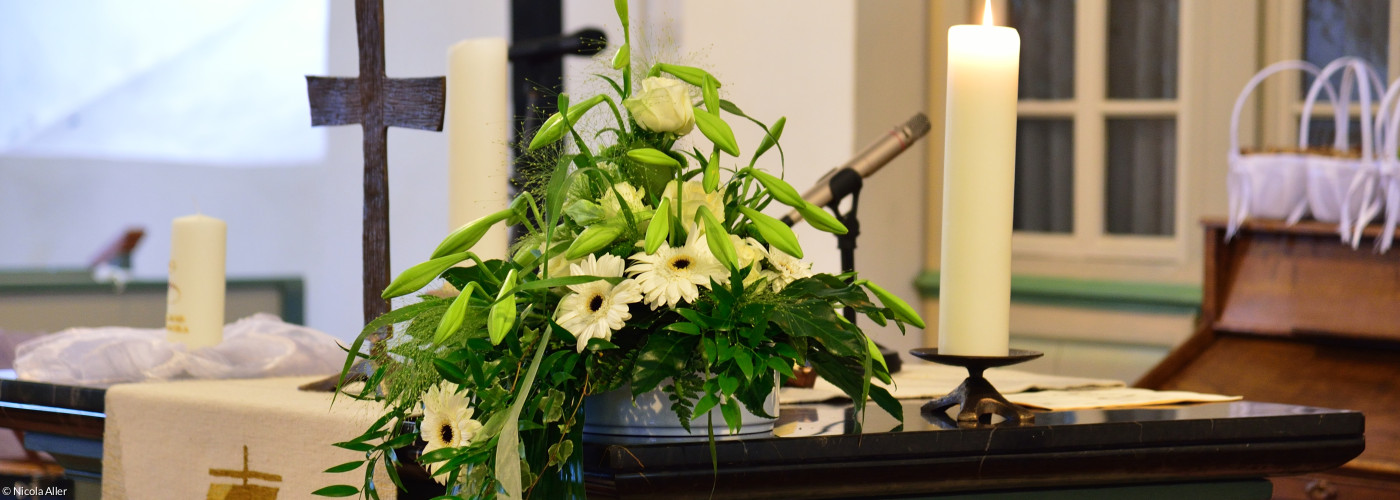 Zu sehen ist ein Altar in einer Dorfkirche mit Kreuz, Blumen, Kerze und Parament mit Schiffmotiv