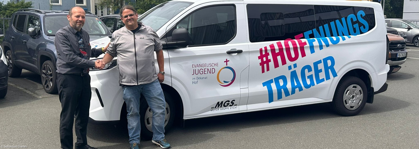 Zwei Männer reichen einander die Hand vor einem weißen Bus mit der Aufschrift "HOFFNUNGSTRÄGER"