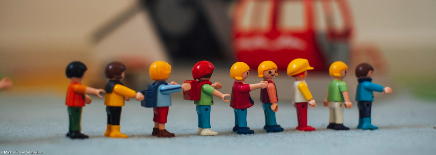 Playmobilmännlein sind in einer Reihe aufgestellt. Im Hintergrund sieht man ein Playmobilfahrzeug