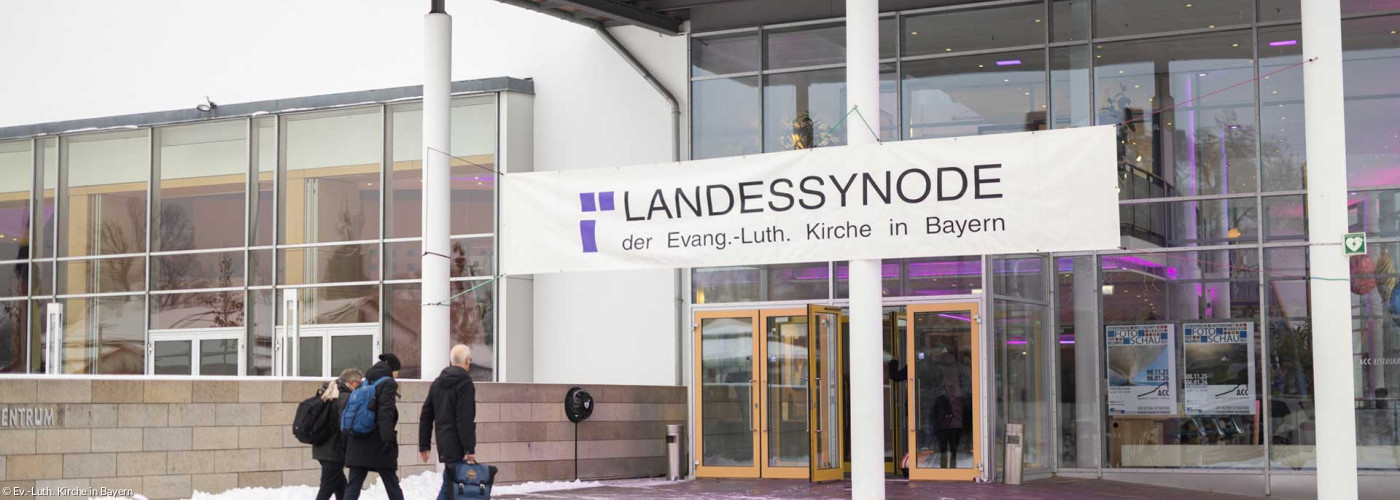 Drei Menschen gehen in ein Kongressgebäude. Oben prangt das Banner "Landessynode"