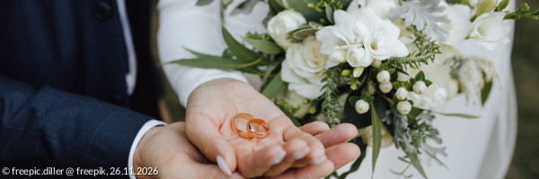 Ein Brautpaar hält Eheringe gemeinsam in der Hand, ein weißer Brautstrauß ist zu sehen
