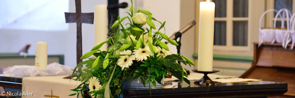 Zu sehen ist ein Altar in einer Dorfkirche mit Kreuz, Blumen, Kerze und Parament mit Schiffmotiv
