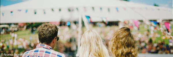 Drei junge Menschen sind von hinten auf einem Festival zu sehen, im Hintergrund ein Partyzelt mit einer Wimpelgirlande