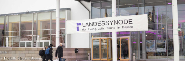 Drei Menschen gehen in ein Kongressgebäude. Oben prangt das Banner "Landessynode"