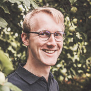 Junger lächelnder Mann mit Poloshirt, blondem Seitenscheitel und Brille vor Blätterhintergrund